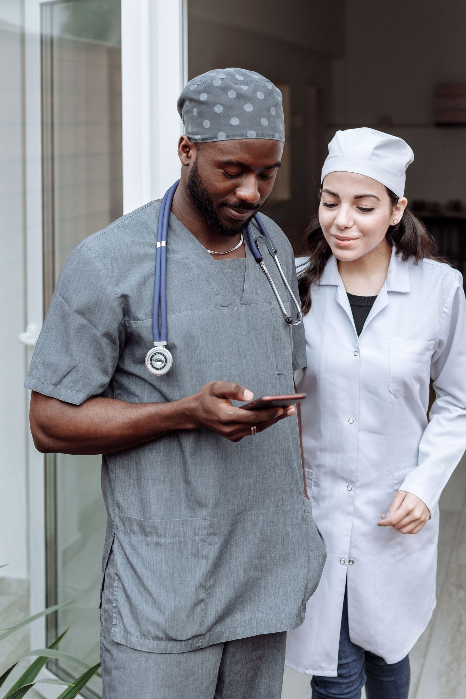 Two healthcare professionals in scrubs checking a phone together outside a home.