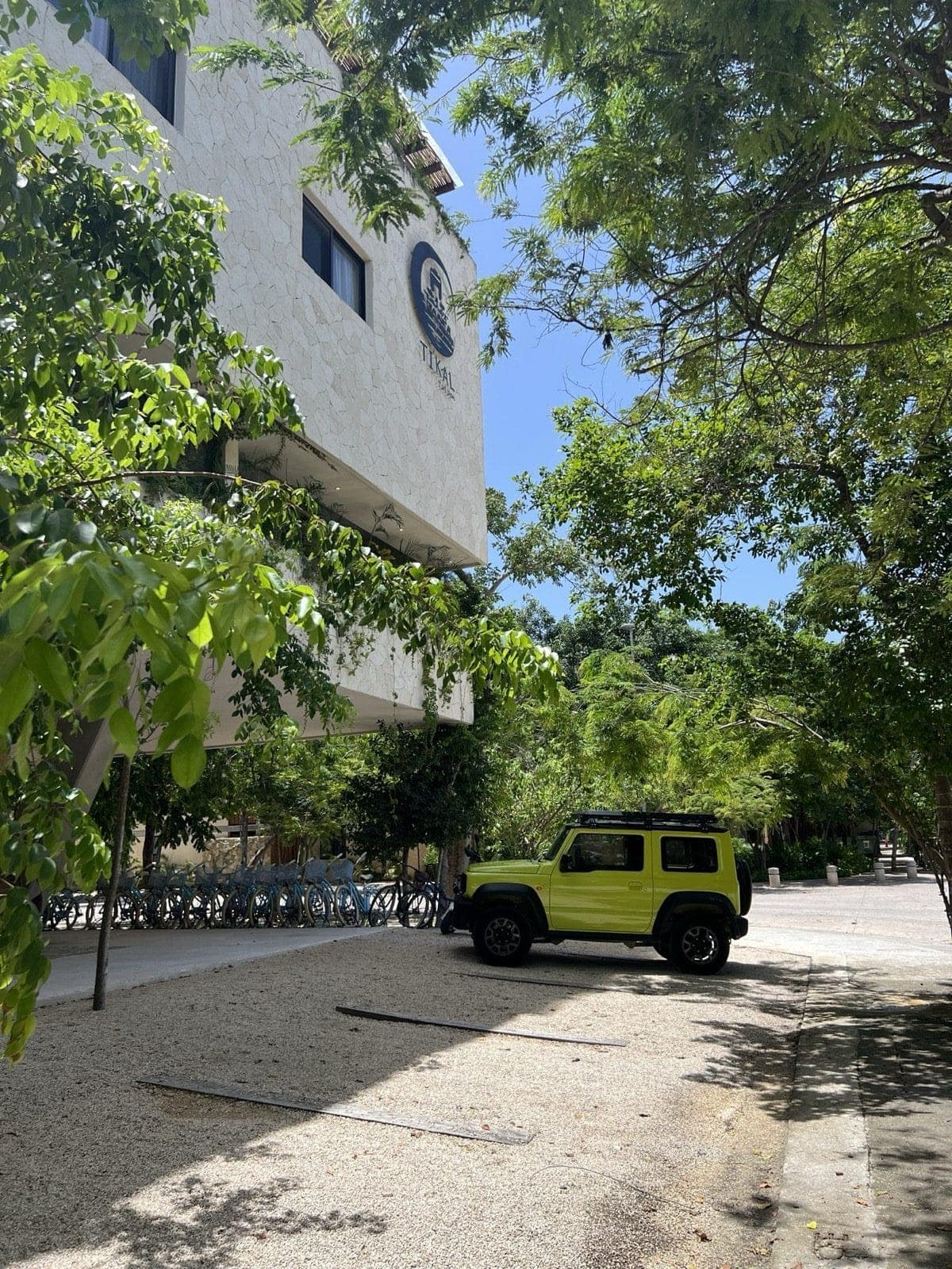 A bright green Jimny parked along a shaded Aldea Zamá street beside a stucco condominium — the neighborhood's everyday rhythm