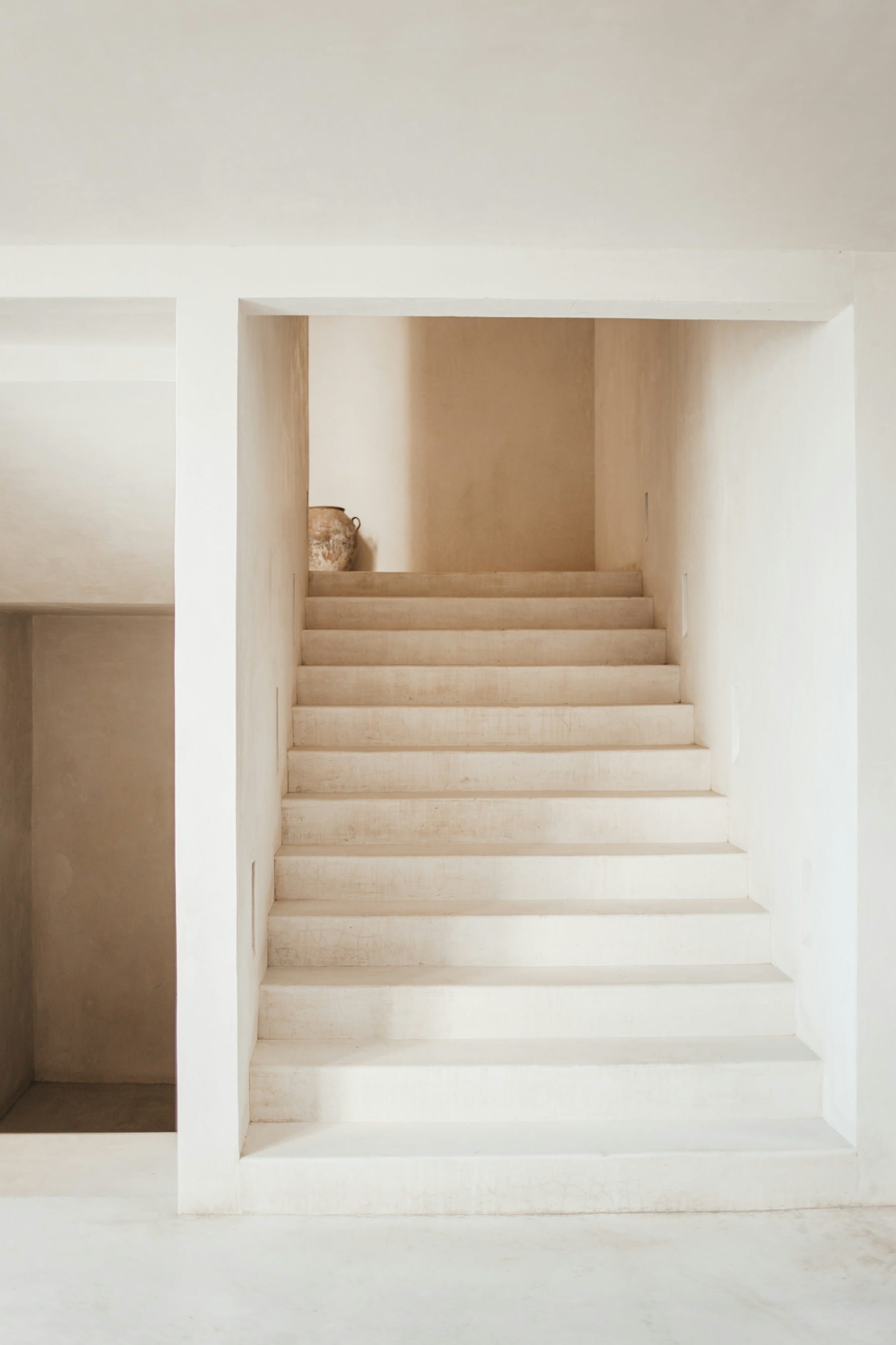 Tulum villa interior design reference — lime-plaster staircase rising to a soft-lit landing, a single terracotta vessel resting at the top